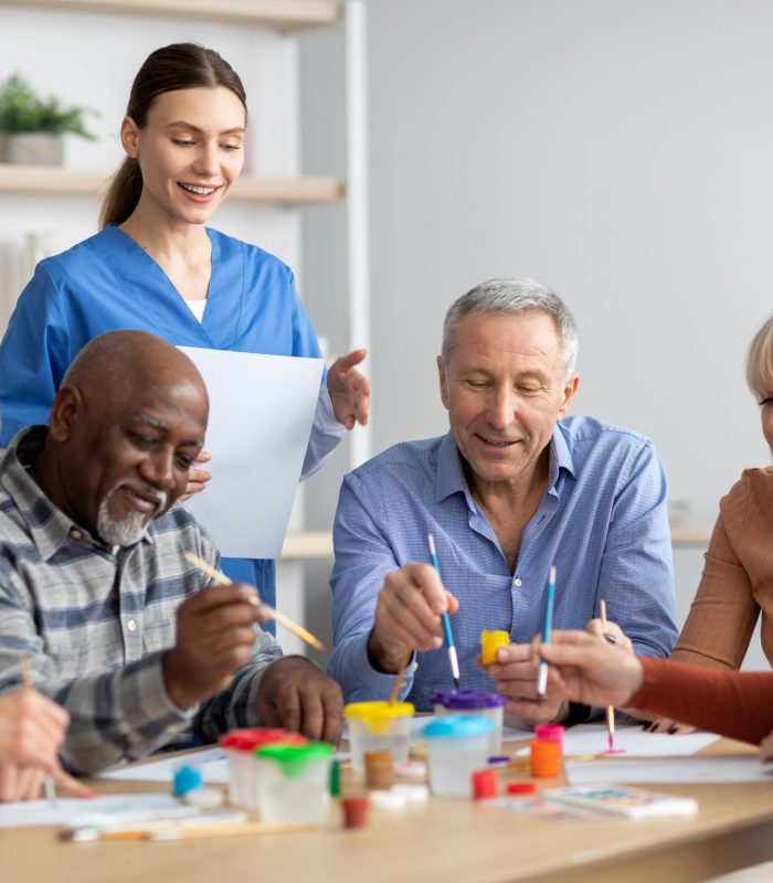 Happy multiracial elderly men and women sitting around table and drawing at nursing home, doing arts and crafts together, young lady nurse assisting group of senior people, copy space