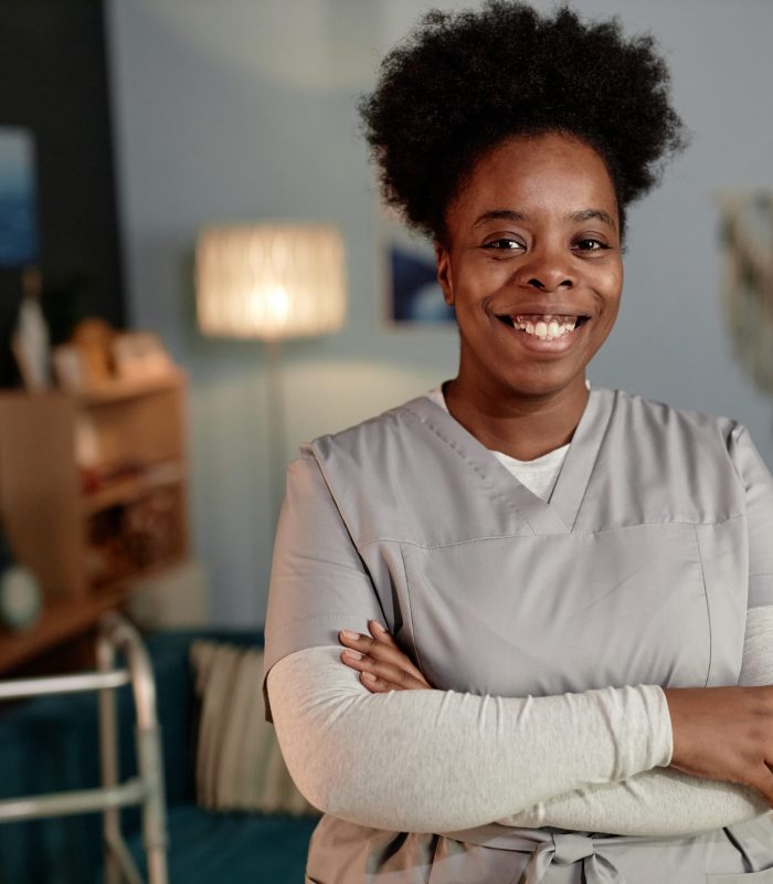 Medium portrait of African American female social worker beaming with smile wearing scrubs posing with arms crossed during house visit to patient, copy space
